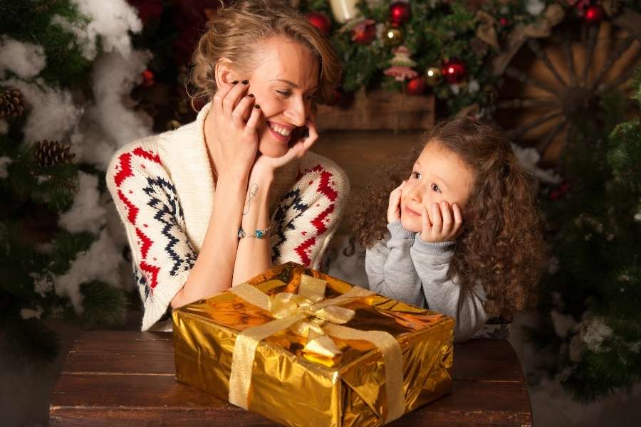 Mère souriante et sa petite fille bouclée admirant ensemble une grande boîte cadeau dorée pour la fête des mères, devant un décor de sapin festif.