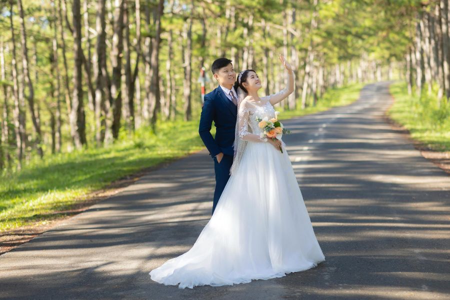 De belles photos de mariage d'un couple posant sur une route bordée d'arbres, la mariée levant la main vers le ciel.