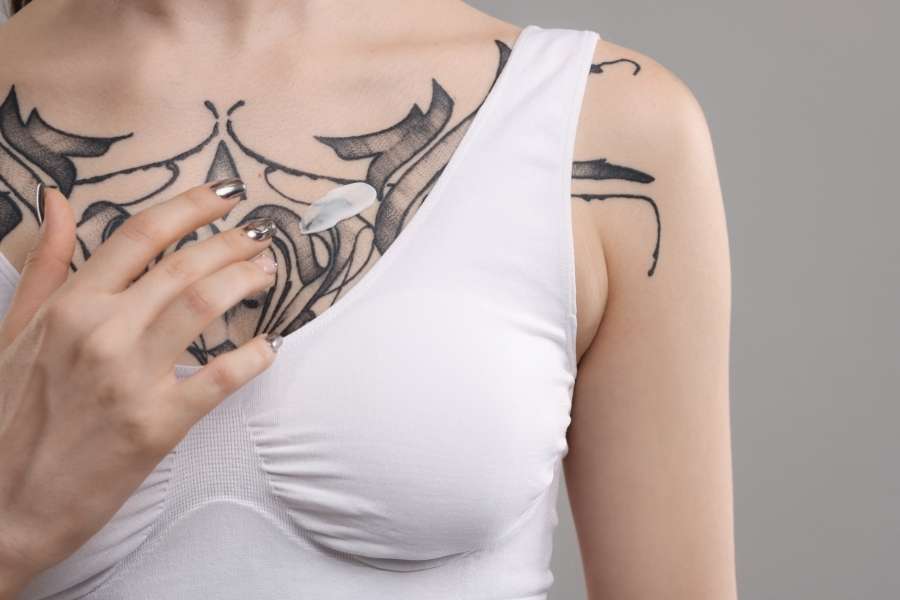 Une personne portant un débardeur blanc applique une fine couche de crème cicatrisante blanche sur son tatouage ornemental noir frais situé sur le sternum et la poitrine.