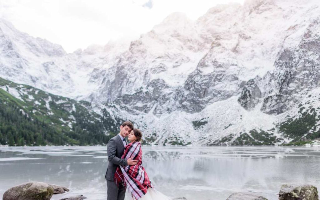 Couple de mariés enlacé sous un plaid chaud au bord d'un lac alpin pour organiser un mariage a la montagne ete hiver.
